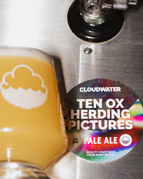 closeup of Cloudwater craft beer, pale ale 'Ten Ox Herding Pictures' poured from tap, bright orangey-yellow with the round keg badge situated next to it