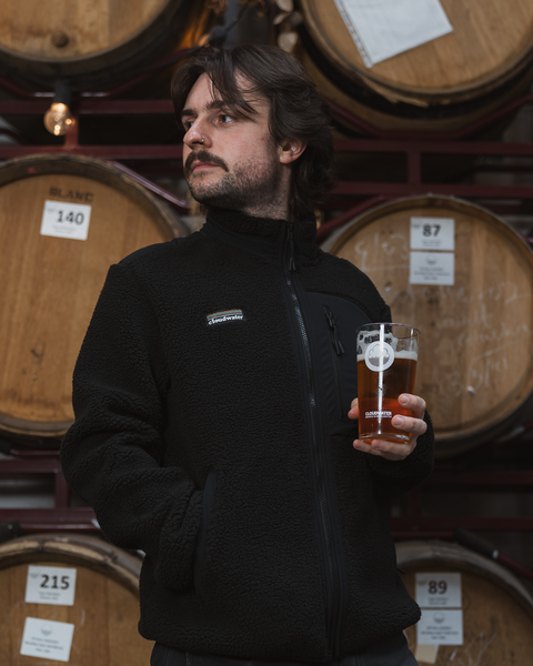 Man wearing a black Cloudwater branded fleece jacket in front of barrels, with a pint of smoked marzen in hand