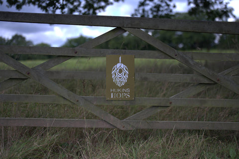 Hukins Hop farm in Kent, fence in a field with the Hukins Hop logo, a hop flower. Cloudwater Brew Co visited Hukins for their 2025 hop harvest and created a fresh hop beer with the Challenger variety