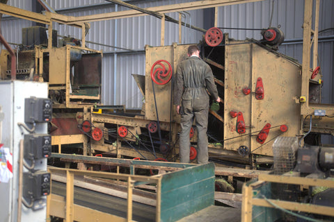 Inside the processing area at Hukins Hop farm in Kent, for UK Hop Harvest 2025 at Hukins Hops. A man in overalls has his back to the camera, facing a yellow box-like piece of machinery with red wheels & pulleys on.