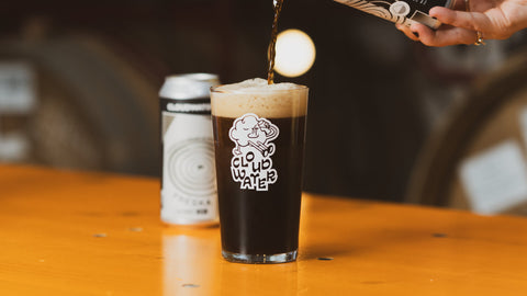 A hand pours dark AF stout beer FRESKA from a black and white can into a clear pint glass with a white 'CLOUDWATER' logo, set on a warm wooden table with a blurred second can in the background.
