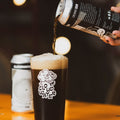 A hand pours dark AF stout beer FRESKA from a black and white can into a clear pint glass with a white 'CLOUDWATER' logo, set on a warm wooden table with a blurred second can in the background.