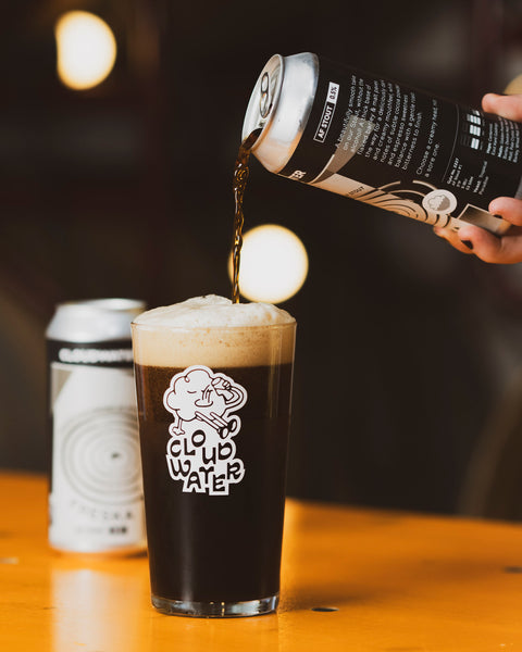 A hand pours dark AF stout beer FRESKA from a black and white can into a clear pint glass with a white 'CLOUDWATER' logo, set on a warm wooden table with a blurred second can in the background.