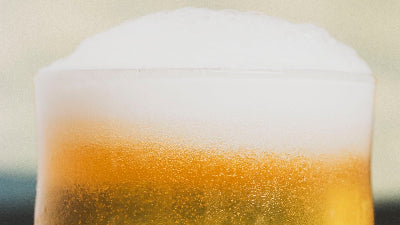 A close-up of a frothy glass of golden XPA by Cloudwater with thick white foam and visible carbonation bubbles, set against a softly blurred warm beige background.