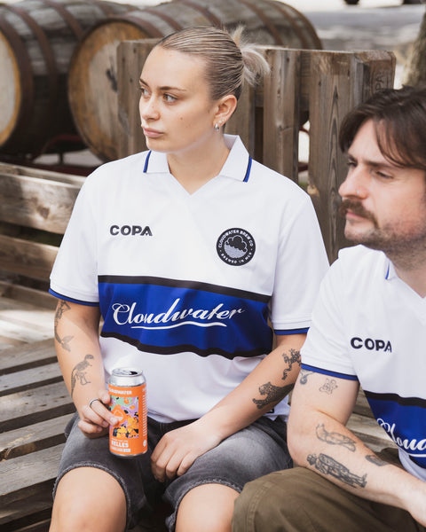 Two people wearing Cloudwater Brew Co x COPA Retro Football Shirts sitting next to wooden barrels.