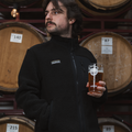 Man wearing a black Cloudwater branded fleece jacket in front of barrels, with a pint of smoked marzen in hand