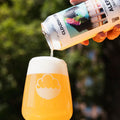 A hand pours Aleph Cloudminer non-alcoholic beer from a white can into a clear stemless glass with a white cloud logo, set against a blurred green outdoor background.
