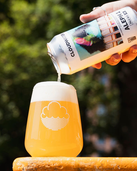 A hand pours Aleph Cloudminer non-alcoholic beer from a white can into a clear stemless glass with a white cloud logo, set against a blurred green outdoor background.