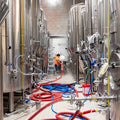 Interior shot of Cloudwater Brewery Production area in their Manchester home. One of the brewers wears orange high vis and is busy working amongst the towering fermentation tanks, with hoses covering the brew floor.