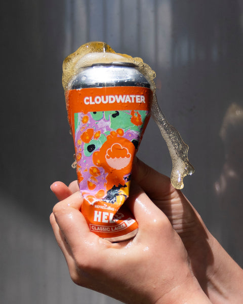 Person crushing a bright orange can of Cloudwater Helles German-Style Beer against a grey background in the sun