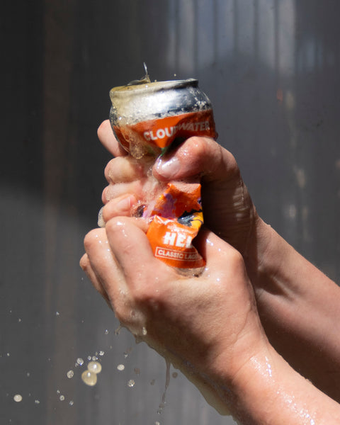 Person crushing a bright orange can of Cloudwater Helles German-Style Beer  against a grey background in the sun