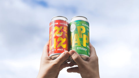 Two hands holding up two aluminum cans of Clouwater Fuzzy & Happy against a cloudy sky background. Cloudwater are celebrating International Beer Day on Friday 7th August