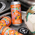 Bright orange patterned cans of Cloudwater Helles classic lager German-Style Beer rests on top of silver kegs beside a football