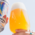 A hand holds a frosted glass filled with golden beer topped with thick white foam, while another hand holds a can of Cloudwater Infinite Space IPA next to it against a pale sky background.