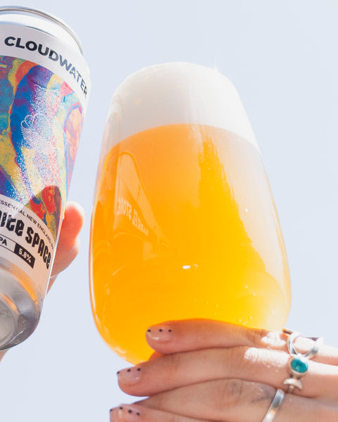A hand holds a frosted glass filled with golden beer topped with thick white foam, while another hand holds a can of Cloudwater Infinite Space IPA next to it against a pale sky background.
