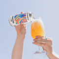 A person holds a can of Cloudwater infinite Space 5% IPA tilted to pour a foamy golden beer into a clear glass, set against a solid light blue sky background.