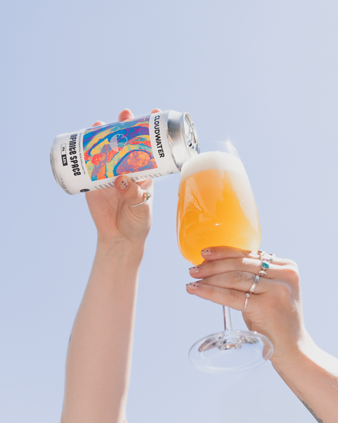 A person holds a can of Cloudwater infinite Space 5% IPA tilted to pour a foamy golden beer into a clear glass, set against a solid light blue sky background.