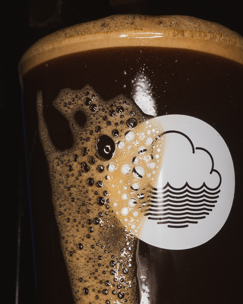 Close-up of a glass of Stollen From Ivor festive stout, with a creamy head and a prominent Cloudwater logo in white on a dark background