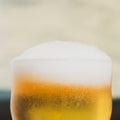 A close-up of a frothy glass of golden XPA by Cloudwater with thick white foam and visible carbonation bubbles, set against a softly blurred warm beige background.