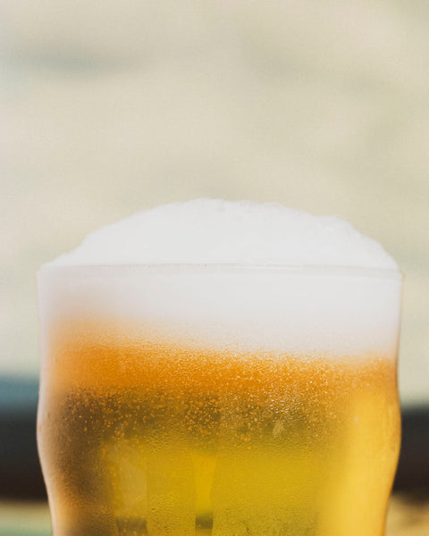 A close-up of a frothy glass of golden XPA by Cloudwater with thick white foam and visible carbonation bubbles, set against a softly blurred warm beige background.