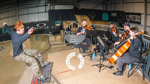 A group of musicians, part of Manchester Camerata, including violinists and a cello player, are seated in a skatepark indoors, playing instruments next to a ramp where a skateboarder is performing. This is the artwork for cloudwater x manchester camerata support your local orchestra pale ale