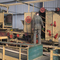 Inside the processing area at Hukins Hop farm in Kent, A man in overalls has his back to the camera, facing a yellow box-like piece of machinery with red wheels & pulleys on. Cloudwater Brew Co visited Hukins for their 2025 hop harvest and created a fresh hop beer with the Challenger variety