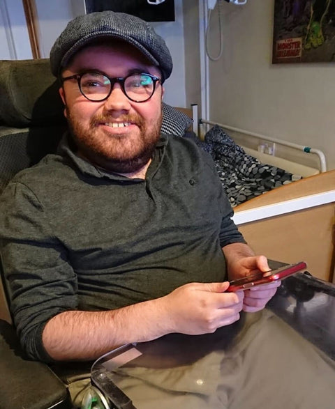 Max Bode living with Muscular Dystrophy, wearing glasses and a cap holding a phone in a hospital room.
