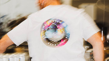 One of Cloudwater's brew team wearing a white t-shirt with a large circular multicolor Cloudwater Logo graphic on the back, features the text 'CLOUDWATER BREWING CO' and 'BREWED IN MCR'