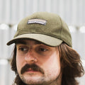 A man wearing a dark olive green wool-blend Cloudwater branded cap. The cap features a black woven outdoor-inspired Cloudwater logo on the front