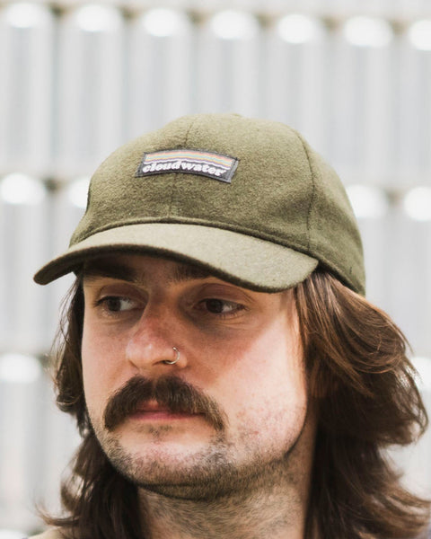 A man wearing a dark olive green wool-blend Cloudwater branded cap. The cap features a black woven outdoor-inspired Cloudwater logo on the front