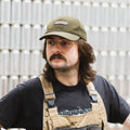 A man wearing a dark olive green wool-blend Cloudwater branded cap. The cap features a black woven outdoor-inspired Cloudwater logo on the front