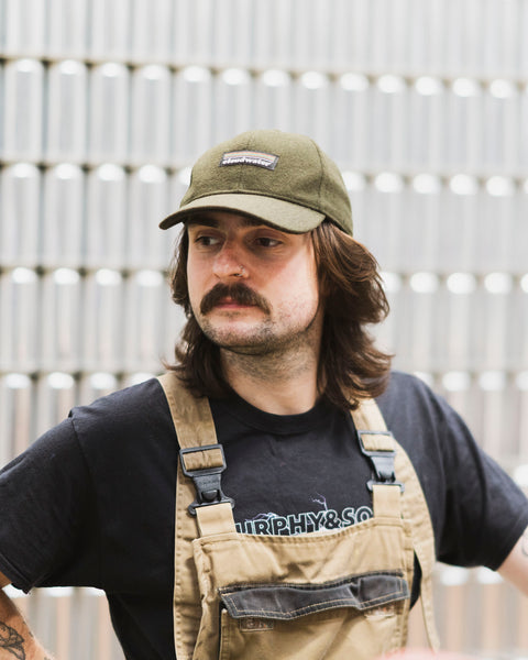 A man wearing a dark olive green wool-blend Cloudwater branded cap. The cap features a black woven outdoor-inspired Cloudwater logo on the front