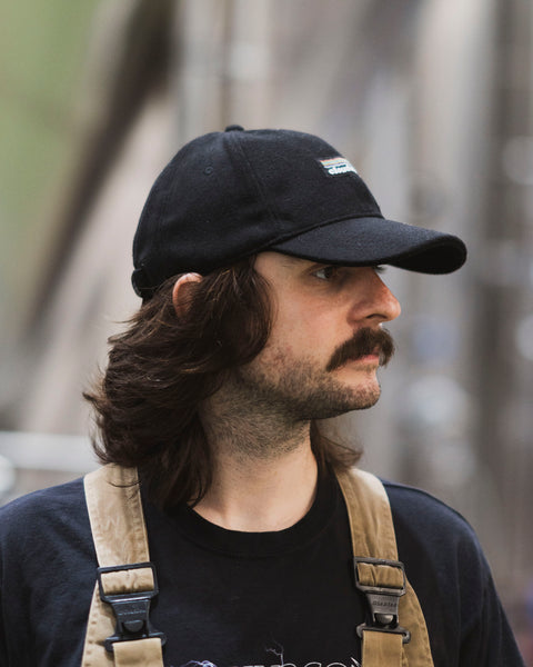 A man wearing a black wool-blend Cloudwater branded cap. The cap features a black woven outdoor-inspired Cloudwater logo on the front