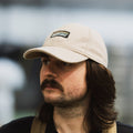A man wearing a stone or cream coloured wool-blend Cloudwater branded cap. The cap features a black woven outdoor-inspired Cloudwater logo on the front