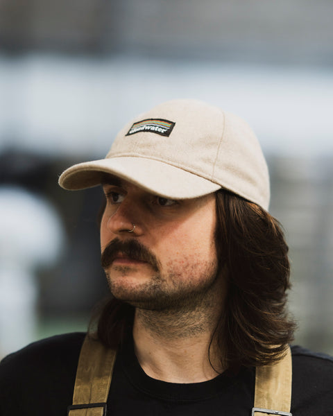 A man wearing a stone or cream coloured wool-blend Cloudwater branded cap. The cap features a black woven outdoor-inspired Cloudwater logo on the front